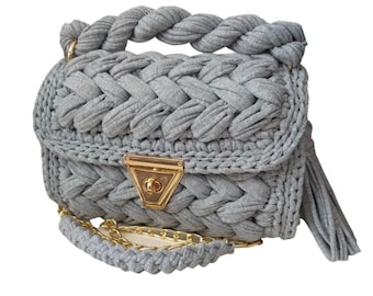 May include: A gray crocheted handbag with a gold chain strap and a gold clasp. The bag has a braided handle and a tassel.