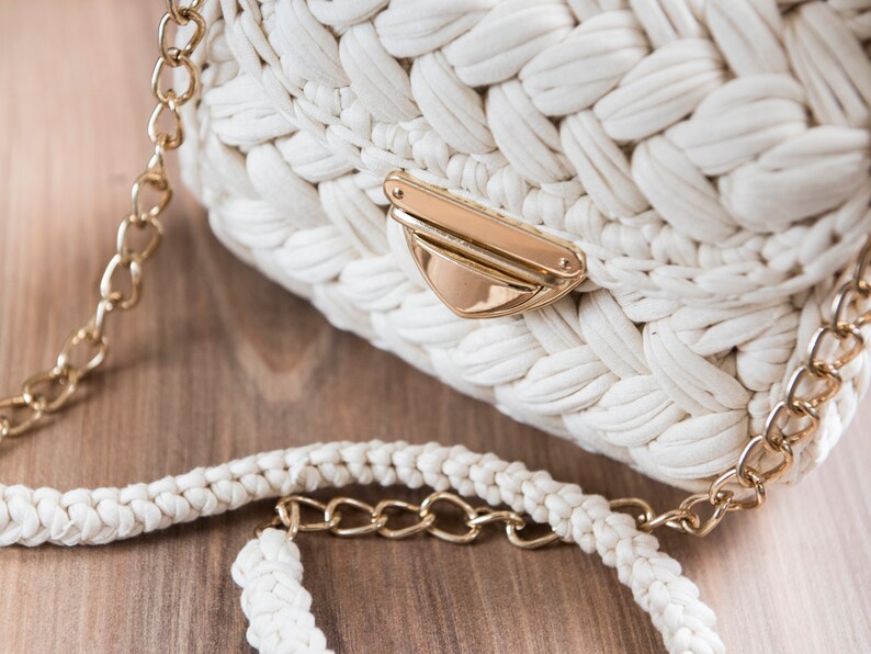 Pu&ograve; includere: Una borsa bianca all'uncinetto con una tracolla a catena dorata e una chiusura a scatto dorata.
