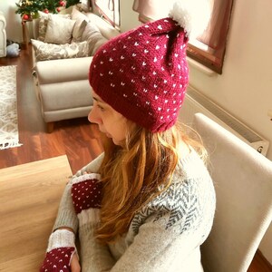 May include: A red knit hat with white hearts and a white pom pom on top. The hat is worn by a person wearing a white sweater with a snowflake pattern.