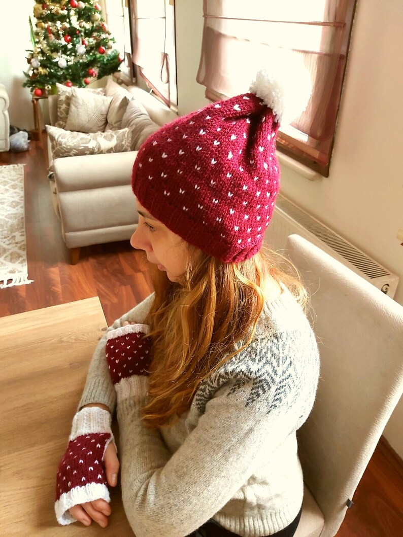 May include: A red knit hat with white hearts and a white pom pom on top. The hat is worn by a person wearing a white sweater with a snowflake pattern.