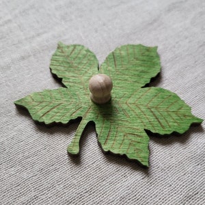 May include: A green wooden leaf with a detailed vein pattern and a small wooden knob in the center.