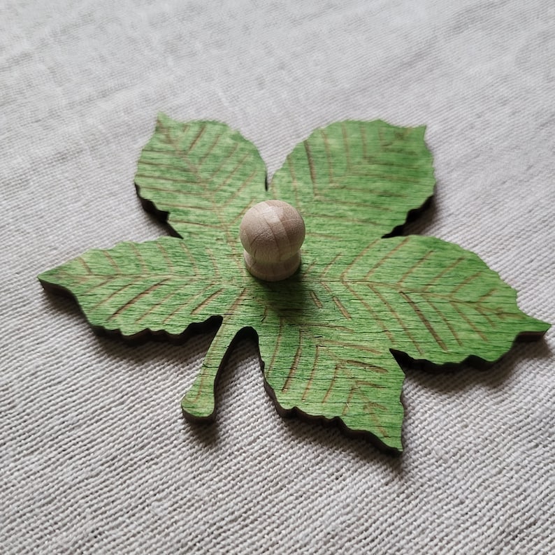 May include: A green wooden leaf with a detailed vein pattern and a small wooden knob in the center.