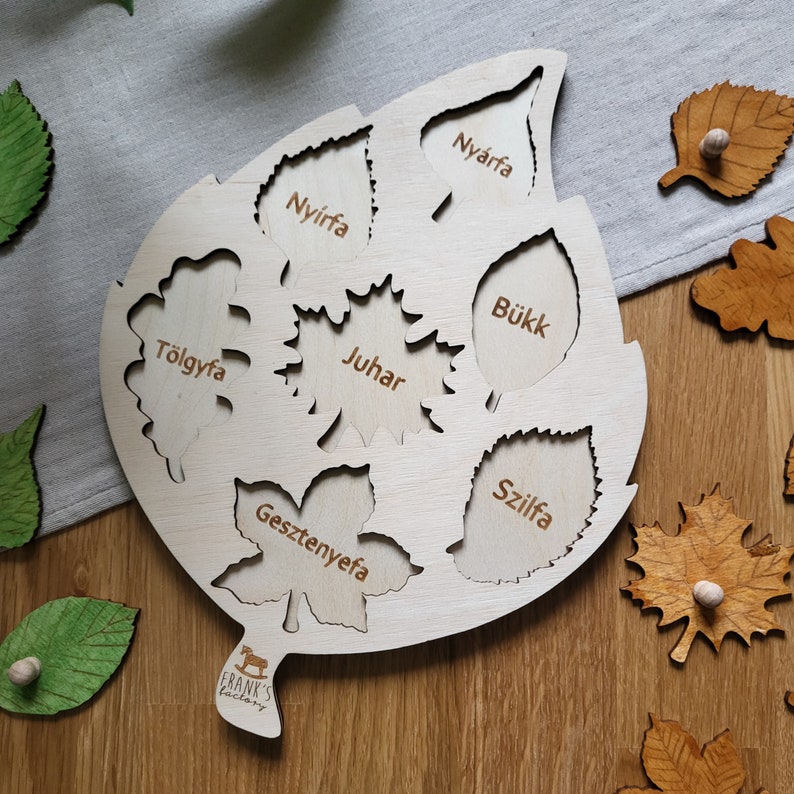 May include: A wooden leaf-shaped puzzle with cutouts of different leaf shapes. Each cutout is labeled with the Hungarian name for the tree the leaf comes from, including "Ny&iacute;rfa", "T&ouml;lgyfa", "Juhar", "B&uuml;kk", "Gesztenyefa", "Szilfa".