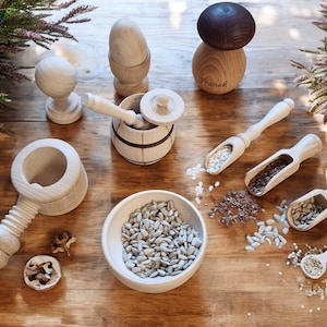 May include: A wooden play set featuring a nutcracker, a barrel, a mushroom, a bowl, and four wooden scoops. The scoops are filled with various seeds, including sunflower seeds, flax seeds, and sesame seeds.