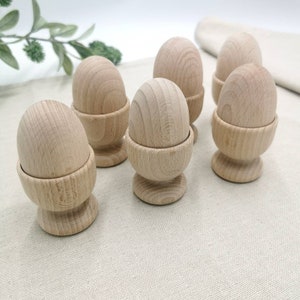 Peut inclure: Six coquetiers en bois clair, chacun contenant un œuf en bois. Les œufs et les coquetiers sont en bois clair, avec des motifs de grain visibles. Les coquetiers ont un design classique avec une base arrondie et un sommet en forme de coupe pour tenir les œufs.