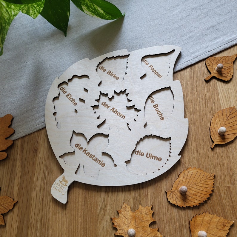 May include: A wooden leaf-shaped puzzle with cutouts of different tree leaves. Each cutout is labeled with the German name for the tree: die Birke, die Eiche, die Pappel, der Ahorn, die Buche, die Kastanie, die Ulme.