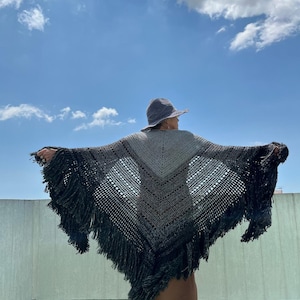 May include: A handmade, open-weave shawl with a fringed edge. The shawl features a gradient of gray and black, with a lighter gray center. The person is wearing a gray hat and a brown dress. The background is a blue sky with clouds.
