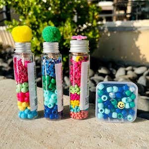 May include: Four small glass jars filled with colorful beads and topped with pom-poms. The jars have labels that say "UMBRELLA & HUGS". A small clear plastic container with blue and green beads is also shown.