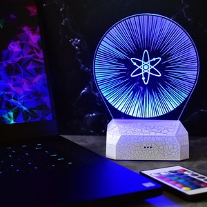May include: A white LED night light with a blue and white atom design. The light is on a white base with a cracked texture. A remote control with color buttons is in the foreground.