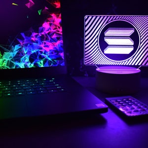 May include: A black laptop with a green, blue, and pink abstract design on the screen. A white LED night light with a white logo on a black background is on a white base. A black remote control is on the table.