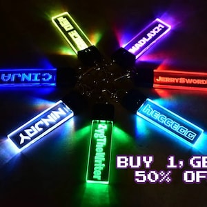 May include: Seven colorful LED keychains with different names on them. The keychains are arranged in a circle and are lit up. The text "BUY 1, GET 1 50% OFF!" is visible at the bottom of the image.