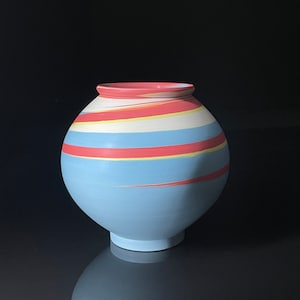 Puede incluir: Un jarrón de cerámica azul y blanco con patrones de espiral rojo y amarillo. El jarrón tiene un cuerpo redondo y una abertura ancha.