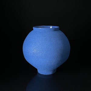 Könnte beinhalten: Eine blaue Keramikvase mit strukturierter Oberfläche. Die Vase hat einen runden Körper und eine breite Öffnung.