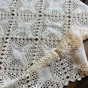 Puede incluir: Un mantel de color blanco roto que presenta un patrón repetitivo de paneles de tela cuadrados con centros florales bordados, unidos por bordes de encaje de ganchillo intrincados. El tejido muestra un delicado diseño calado en toda la pieza.