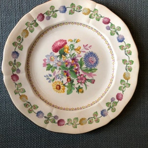 Puede incluir: Un plato de cena de cerámica blanca con un diseño floral. El plato tiene un borde festoneado y un borde dorado. El diseño floral presenta flores rosas, moradas, amarillas y azules con hojas verdes.