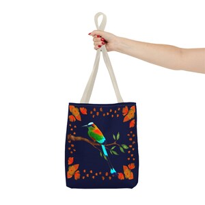 May include: A navy blue tote bag featuring a colorful bird perched on a branch, surrounded by orange butterflies. The bag has cream-colored handles and is held up by a person's hand.