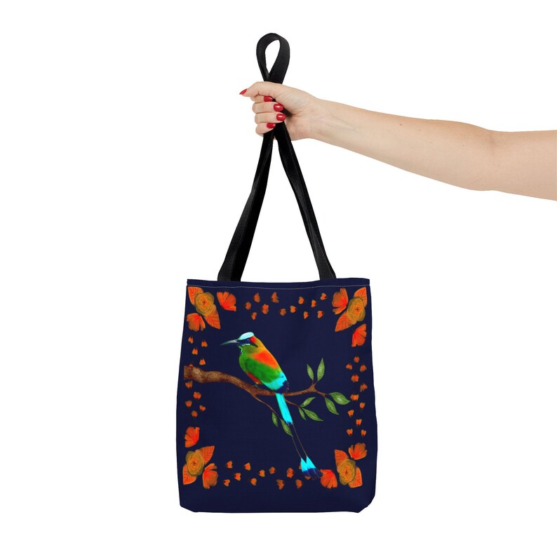 May include: A navy blue tote bag featuring a colorful bird perched on a branch, surrounded by orange butterflies. The bag has black handles and is held up by a person.