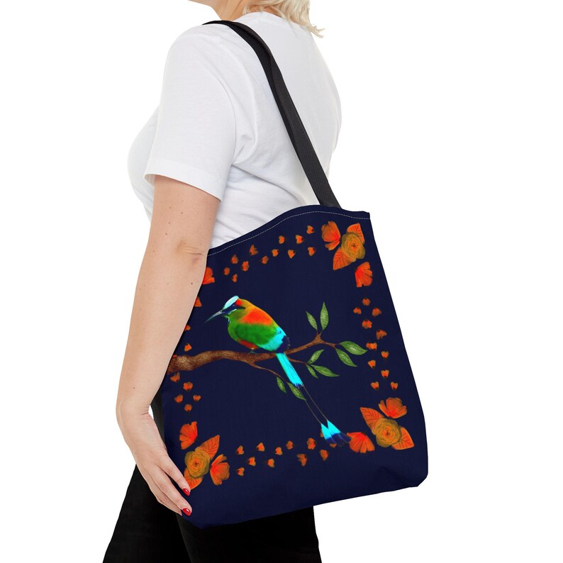 May include: A navy blue tote bag featuring a colorful bird perched on a branch. The bird has green, red, and blue feathers. Orange floral accents and a black strap complete the design. The bag is carried over the shoulder.