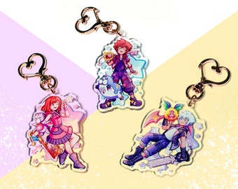 May include: Three acrylic keychains featuring colorful anime-style characters. Each keychain has a gold-colored heart-shaped clasp. The characters are depicted with vibrant colors and are set against a pastel background with star accents.