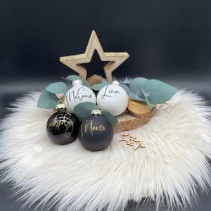 May include: A festive arrangement featuring personalized Christmas ornaments. The ornaments are black and white with gold lettering, displaying names like "Melanie" and "Marco." A wooden star and eucalyptus sprigs add to the holiday theme, set on a fluffy white surface.