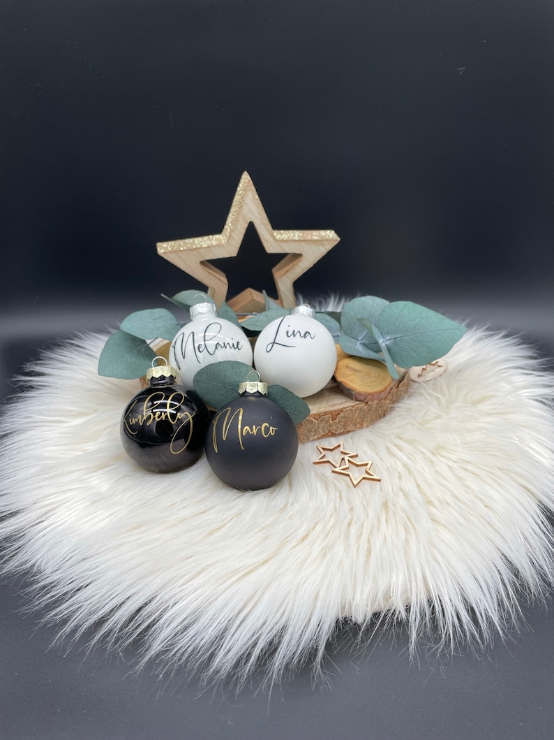 May include: A festive arrangement featuring personalized Christmas ornaments. The ornaments are black and white with gold lettering, displaying names like "Melanie" and "Marco." A wooden star and eucalyptus sprigs add to the holiday theme, set on a fluffy white surface.