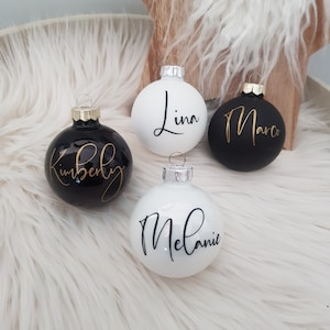May include: Four personalized Christmas ornaments. Two black ornaments with gold lettering that read "Kimberly" and "Marco". Two white ornaments with black lettering that read "Lina" and "Melanie". Each ornament has a silver or gold cap.