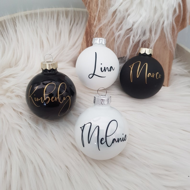 May include: Four personalized Christmas ornaments. Two black ornaments with gold lettering that read "Kimberly" and "Marco". Two white ornaments with black lettering that read "Lina" and "Melanie". Each ornament has a silver or gold cap.