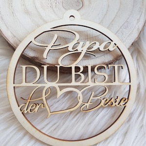 May include: A wooden ornament with the text "Papa du bist der Beste" in a heart shape. The ornament is light brown and has a hole at the top for hanging.