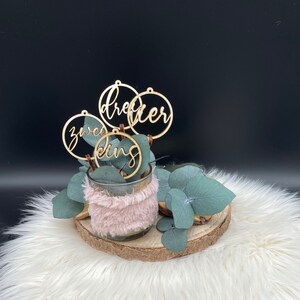 May include: A decorative arrangement featuring a glass vase wrapped in pink faux fur, filled with eucalyptus leaves and wooden ring ornaments. The ornaments spell out the words "dreier", "zweed", and "king". The arrangement sits on a wooden slice and white fur.
