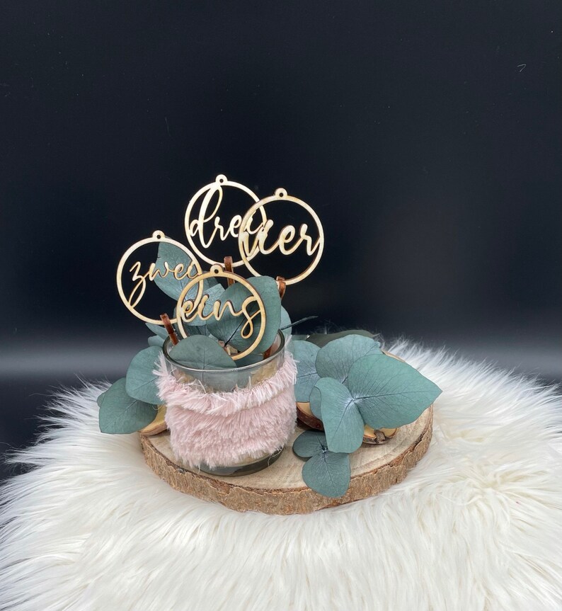 May include: A decorative arrangement featuring a glass vase wrapped in pink faux fur, filled with eucalyptus leaves and wooden ring ornaments. The ornaments spell out the words "dreier", "zweed", and "king". The arrangement sits on a wooden slice and white fur.