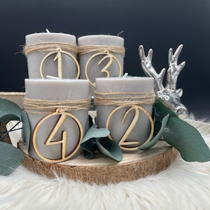 May include: Four gray pillar candles with wooden numbers 1, 2, 3, and 4, wrapped with twine. The candles are on a wooden slice with faux greenery and a silver deer figurine. The background is dark.