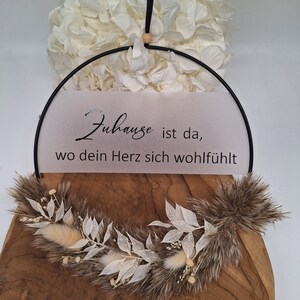 Könnte beinhalten: Ein schwarzer Metallring mit einem weißen Papiereinsatz, auf dem "Zuhause ist da, wo dein Herz sich wohlfühlt" steht. Der Ring ist mit einem braunen und weißen Blumenkranz verziert.