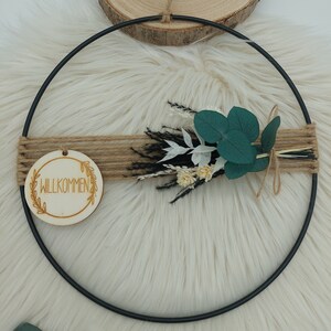 May include: A black metal hoop wreath with a wooden sign that says "Willkommen" and a bouquet of dried flowers with green leaves.
