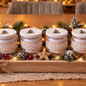 May include: Four glass jars with white lids, each labeled 1. ADVENT through 4. ADVENT, sit in a wooden tray. The jars are surrounded by pine branches, pinecones, red ornaments, wooden star cutouts, and a small light-up house and tree.