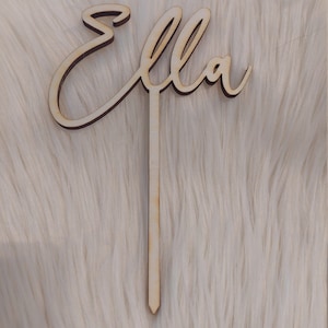 Könnte beinhalten: Ein Holz-Kuchentopper mit dem Namen "Ella" in Kursivschrift. Der Topper ist an einem dünnen Holzstab befestigt.