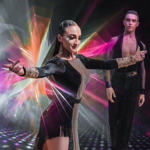 Può includere: Una donna con un abito nero e argento con paillettes e un orlo con frange sta ballando con un uomo con una camicia e pantaloni neri. Entrambi sono in uno studio ben illuminato con luci colorate e un pavimento a motivi.
