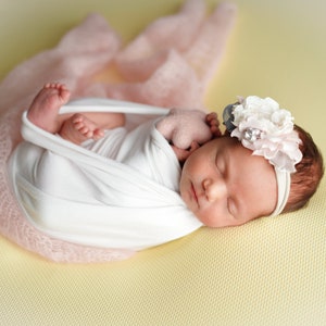 Op de afbeelding: Een pasgeboren babymeisje slaapt rustig in een roze swaddle, met een wit hoofdband met een bloemenaccent. De baby ligt op een gele achtergrond.
