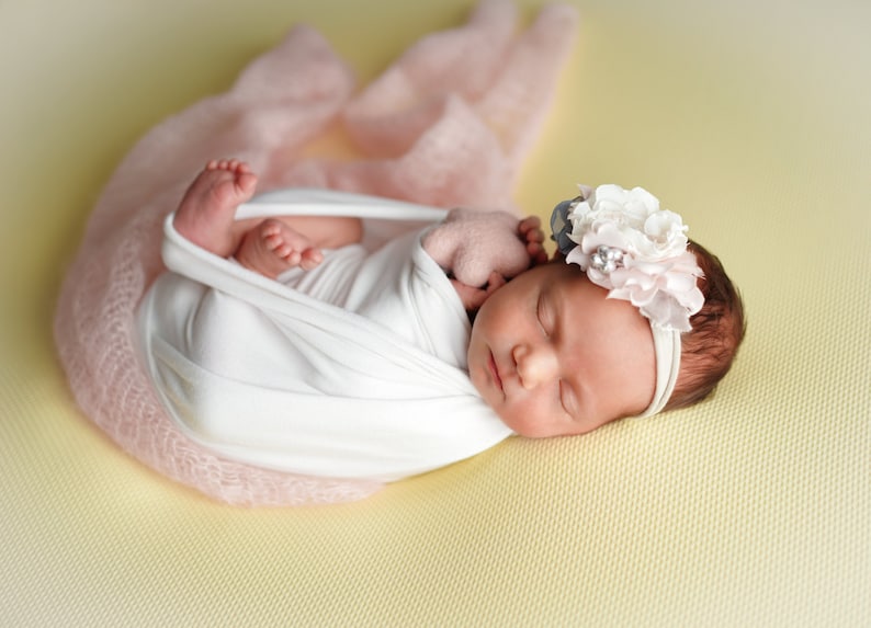 Op de afbeelding: Een pasgeboren babymeisje slaapt rustig in een roze swaddle, met een wit hoofdband met een bloemenaccent. De baby ligt op een gele achtergrond.