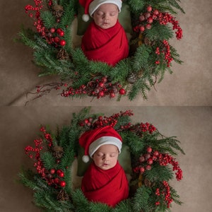 Op de afbeelding: Een pasgeboren baby die een rode kerstmanmuts en een rode swaddle draagt, slaapt in een groene kerstkrans versierd met rode bessen.