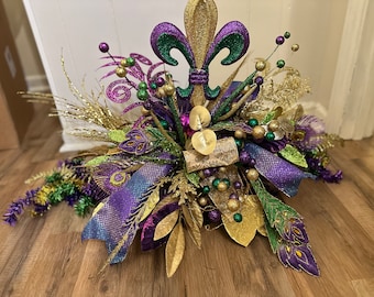May include: A Mardi Gras centerpiece featuring a gold, green, and purple fleur-de-lis, surrounded by peacock feathers, gold glitter, and purple and green beads.