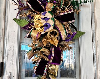 May include: A Mardi Gras themed door wreath with gold, purple, and green accents. The wreath features a gold and purple mask, gold glitter ribbon, and green leaves.