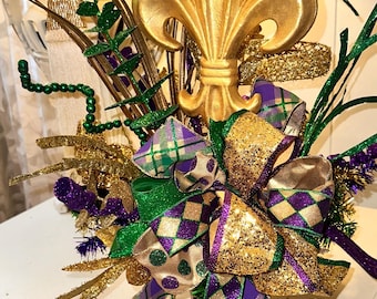 May include: A gold fleur-de-lis centerpiece with a green, purple, and gold bow. The centerpiece is decorated with green and gold glitter and purple and green plaid ribbon.