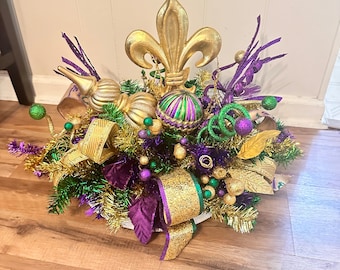 May include: A Mardi Gras centerpiece with a gold fleur-de-lis, purple, green, and gold beads, and a gold and green ornament.