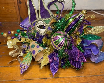 May include: A Mardi Gras themed centerpiece featuring a purple, green, and gold jester mask, a green and gold ornament, and purple and green foliage.