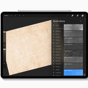 Puede incluir: Una tableta digital que muestra la aplicaci&oacute;n Procreate con la "Biblioteca de pinceles" abierta. La pantalla muestra varias texturas de papel, incluyendo "Paper Texture 16" resaltada en azul. Un l&aacute;piz digital descansa en el borde de la tableta. Fondo blanco.