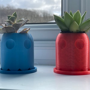 Può includere: Due vasi a forma di fantasma stampati in 3D, uno blu e uno rosso, con piante grasse che crescono all'interno.