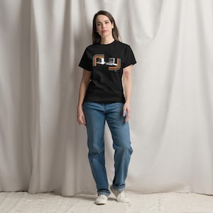 Op de afbeelding: Een zwart T-shirt met korte mouwen en een grafische print van twee vintage desktopcomputers in het midden op de borst, omlijst door horizontale oranje, gele en witte gebogen lijnen voor een retro-geïnspireerd ontwerp.
