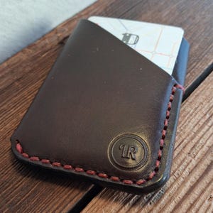 Puede incluir: Un tarjetero de cuero marrón oscuro con costuras rojas. El tarjetero tiene una esquina redondeada y un logotipo "1R" en relieve. Una tarjeta con un diseño de mapa es parcialmente visible. Sobre una superficie de madera.
