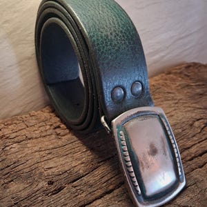 May include: A rolled, dark teal leather belt with a silver-colored rectangular buckle. The belt has two rivets near the buckle. The buckle has a textured border and a brushed metal appearance. The belt is displayed on a weathered wooden surface.