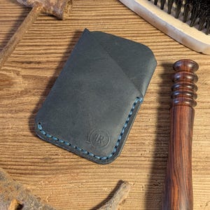 May include: A dark blue leather cardholder with blue stitching and a circular logo. The cardholder is on a wooden surface with a horseshoe, hammer, brush, and a wooden tool.